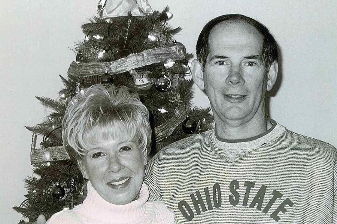 A wife and husband stand beside each other in front of a Christmas tree